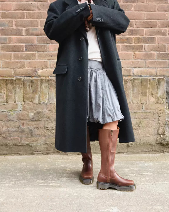 Person in a long coat and brown boots standing against a brick wall.