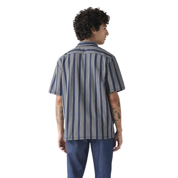 Person in a striped shirt and jeans from behind, with visible tattoos on arms.