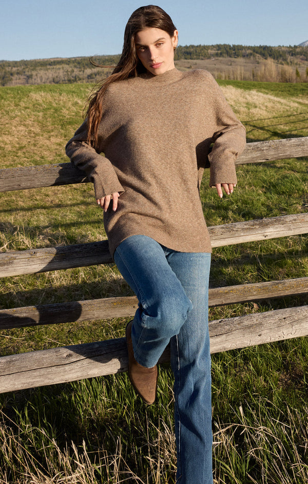 Person in a brown sweater and jeans leaning on a wooden fence in a field.